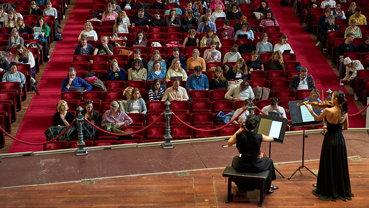 Students studying with laptops in red velvet seats while musicians perform at Amsterdam Concertgebouw