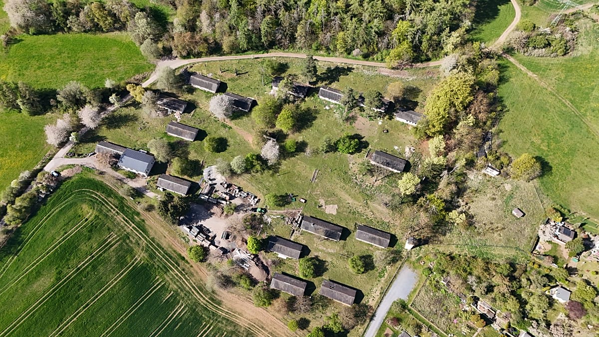 Aerial view of 15 single-story buildings arranged in village formation in German countryside near forest