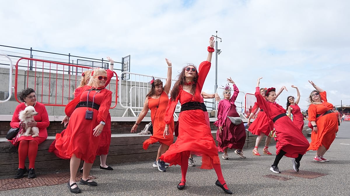** Crowd of people in red dresses dancing together at Folkestone Harbour recreating Kate Bush choreography