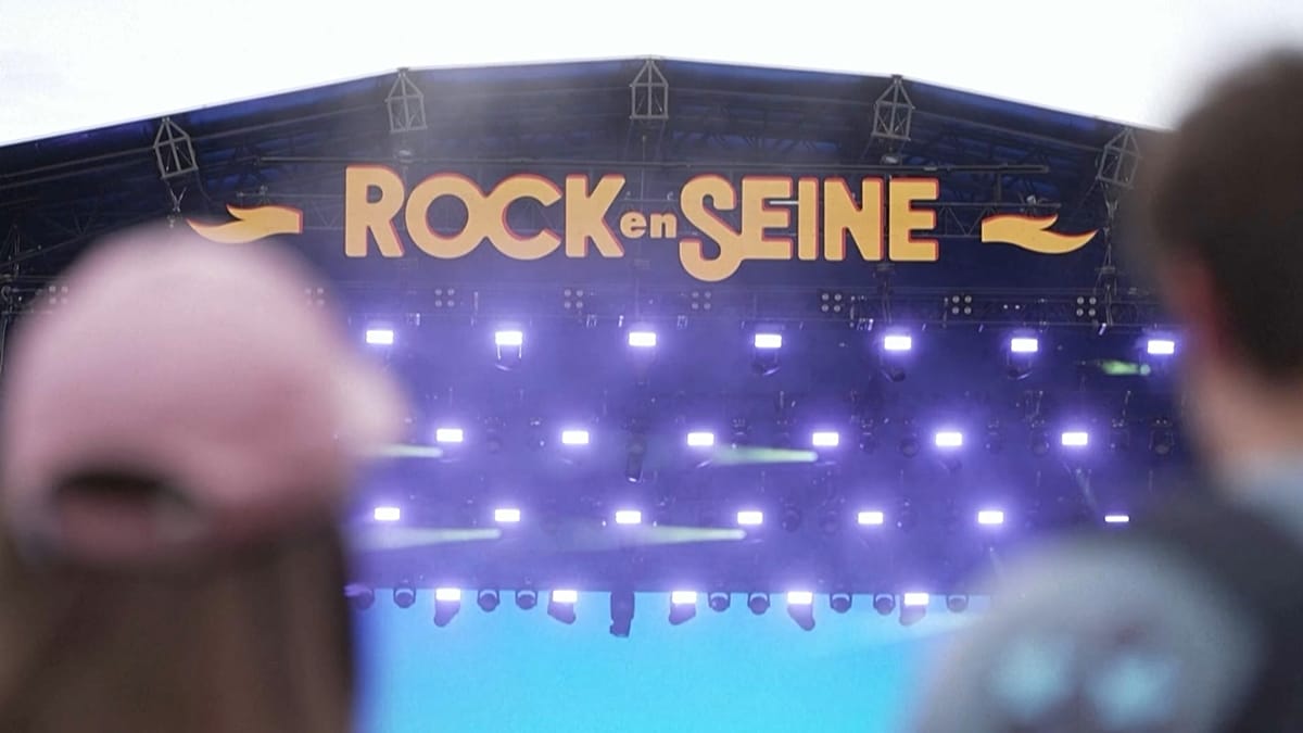 Voice actor wearing headphones narrates concert from festival platform with stage lights visible behind