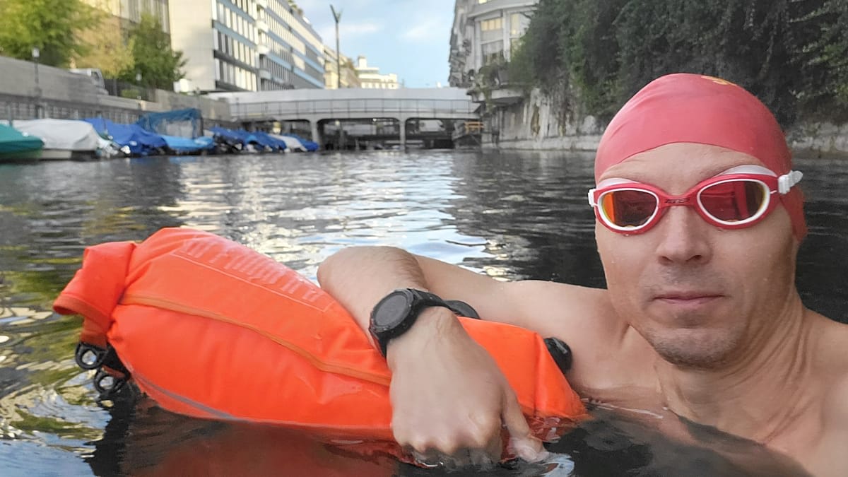 Man swimming in urban canal with waterproof bag, historic buildings lining waterway