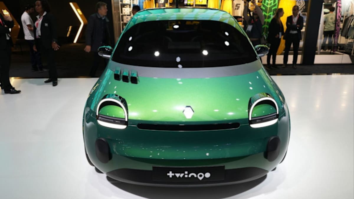 ** Compact electric car charging at station with city street background showing affordable transportation future
