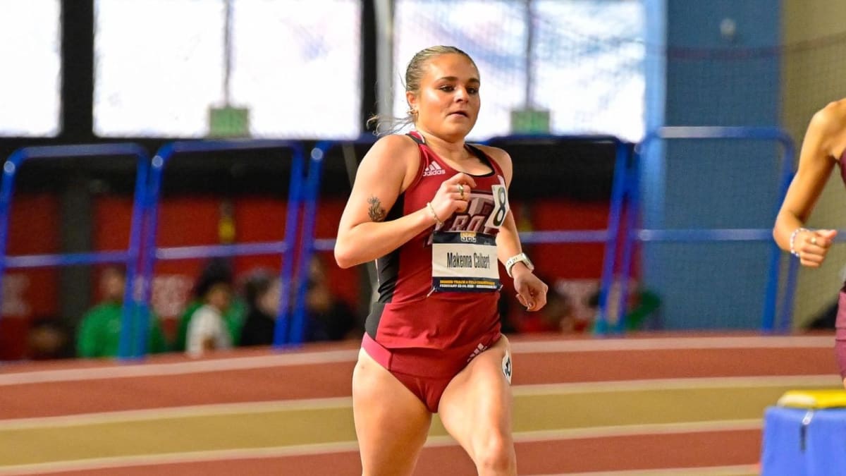 Female college cross country runner competing on outdoor course wearing Troy University uniform