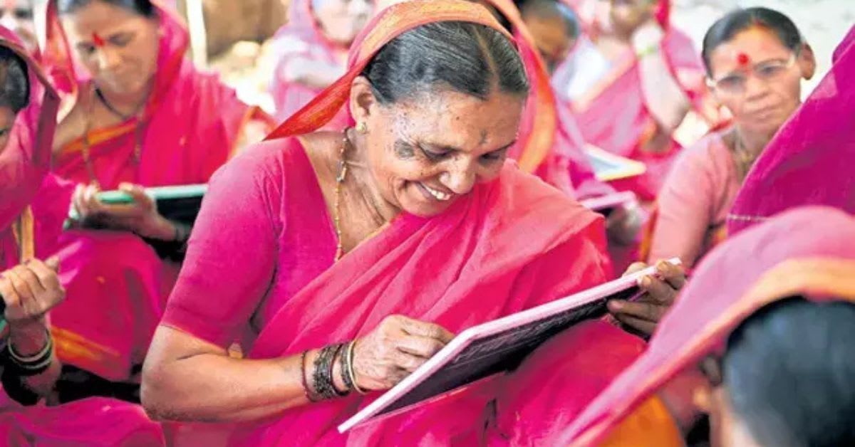 Elderly Indian women in bright pink sarees sitting in classroom holding slates and learning to write