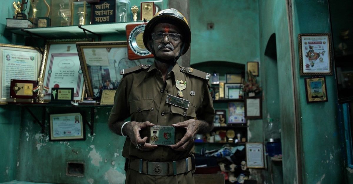 Elderly Indian man in firefighter uniform standing amid newspaper clippings documenting his rescue work