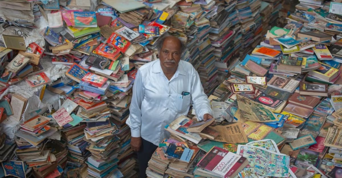 Elderly man Anke Gowda standing among towering shelves filled with thousands of books in his free library