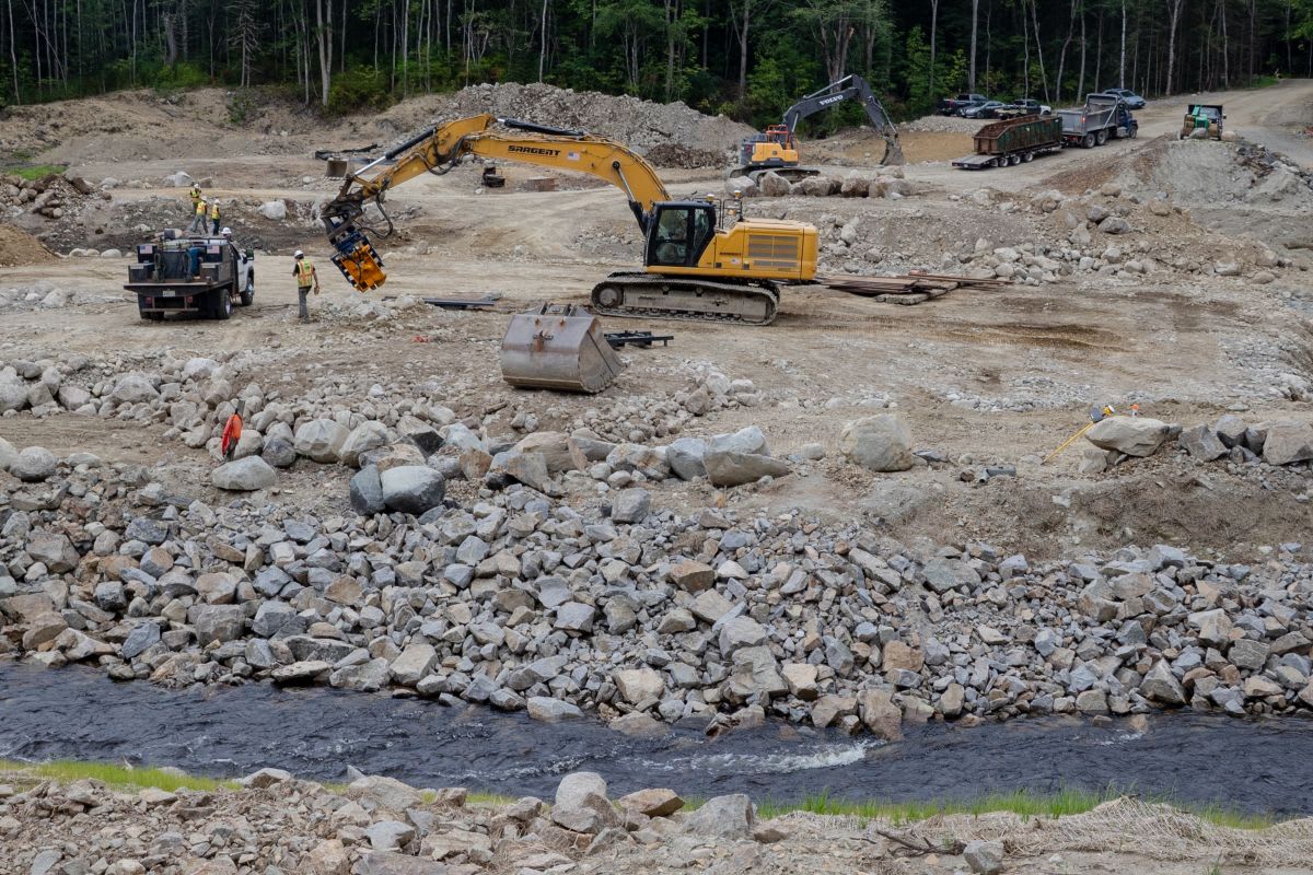 ** Young woman in outdoor work clothes stands near river restoration construction site in Maine