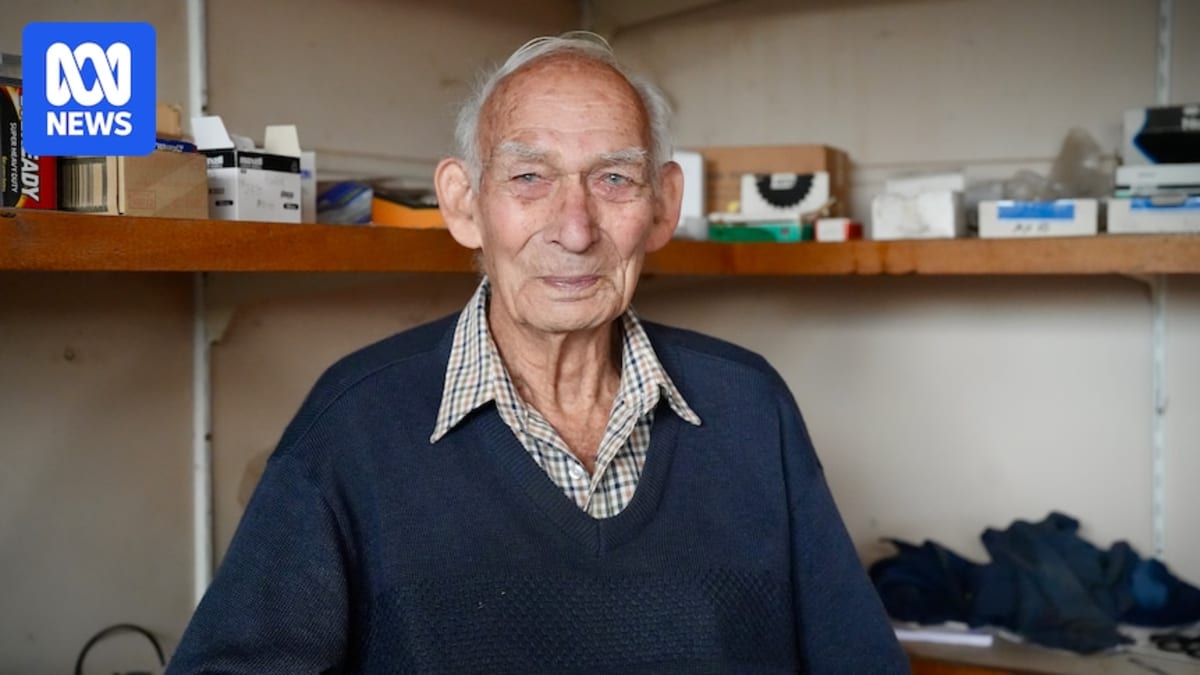 ** Ray Gloster, 99, stands in his family's automotive workshop wearing work clothes and smiling