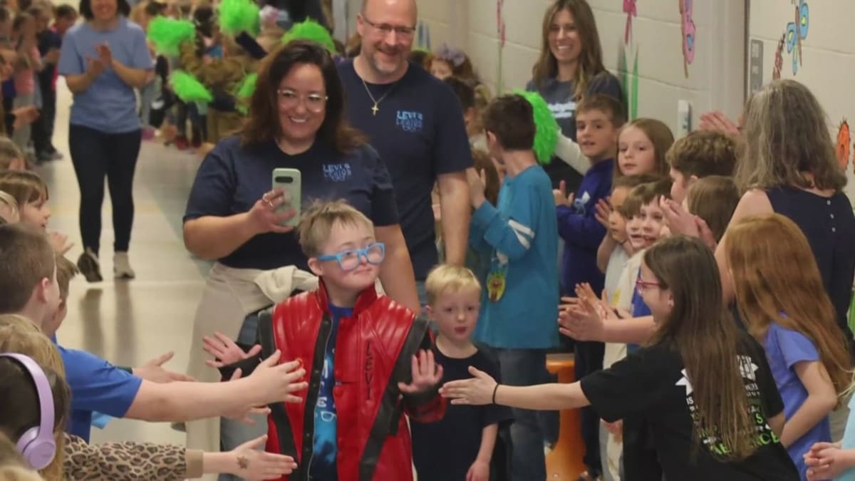 Young boy with Down syndrome smiling in school hallway surrounded by cheering classmates dressed in blue