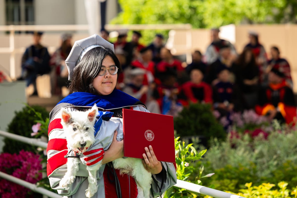Michelle Quien in doctoral robes holds her small dog Vinny wearing matching graduation gown at MIT ceremony