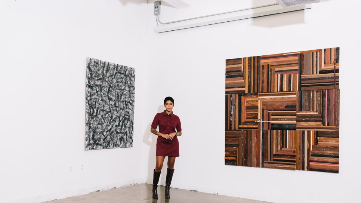 Artist Lex Marie standing beside canvas artwork featuring arranged belts in various skin tones