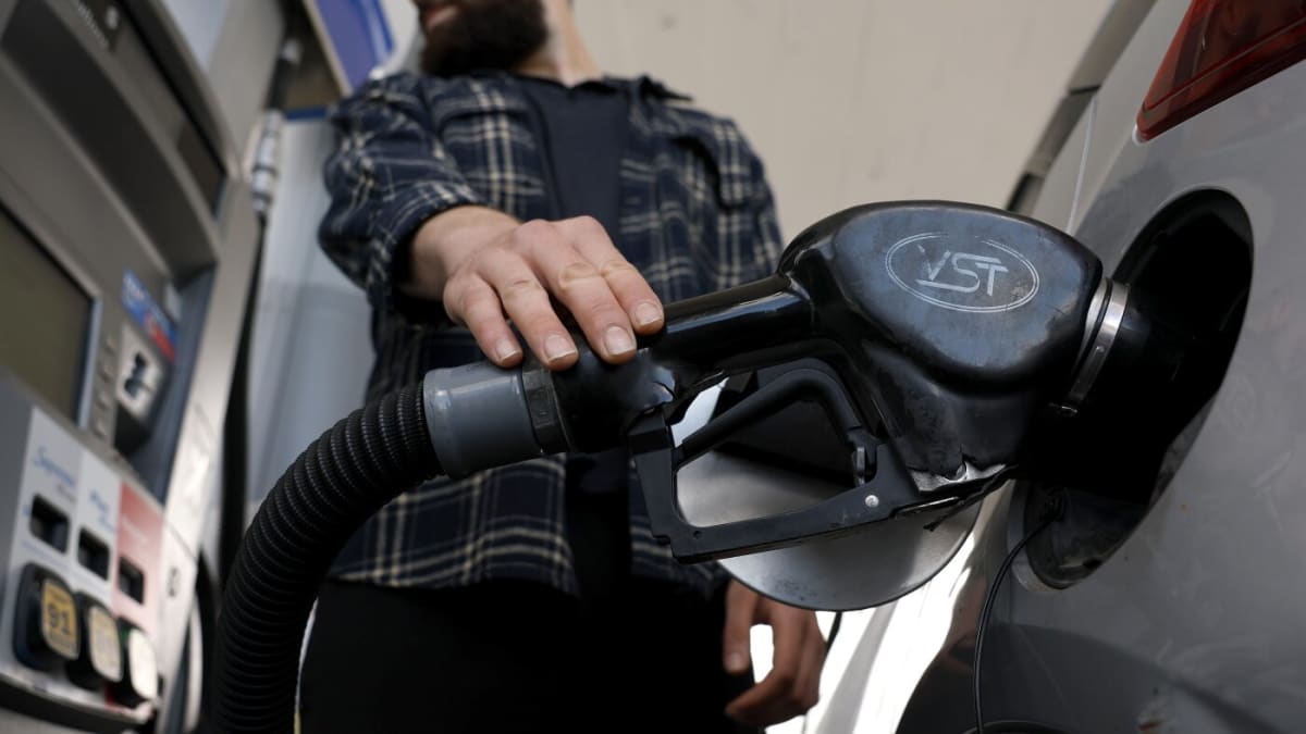 Driver filling electric vehicle at charging station with traditional gas station visible in background