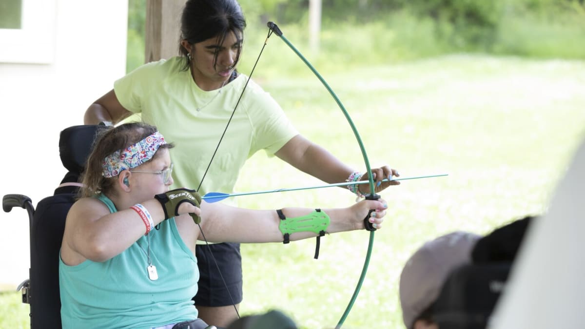 Young camper in wheelchair using hoist system to climb tall rock wall at summer camp