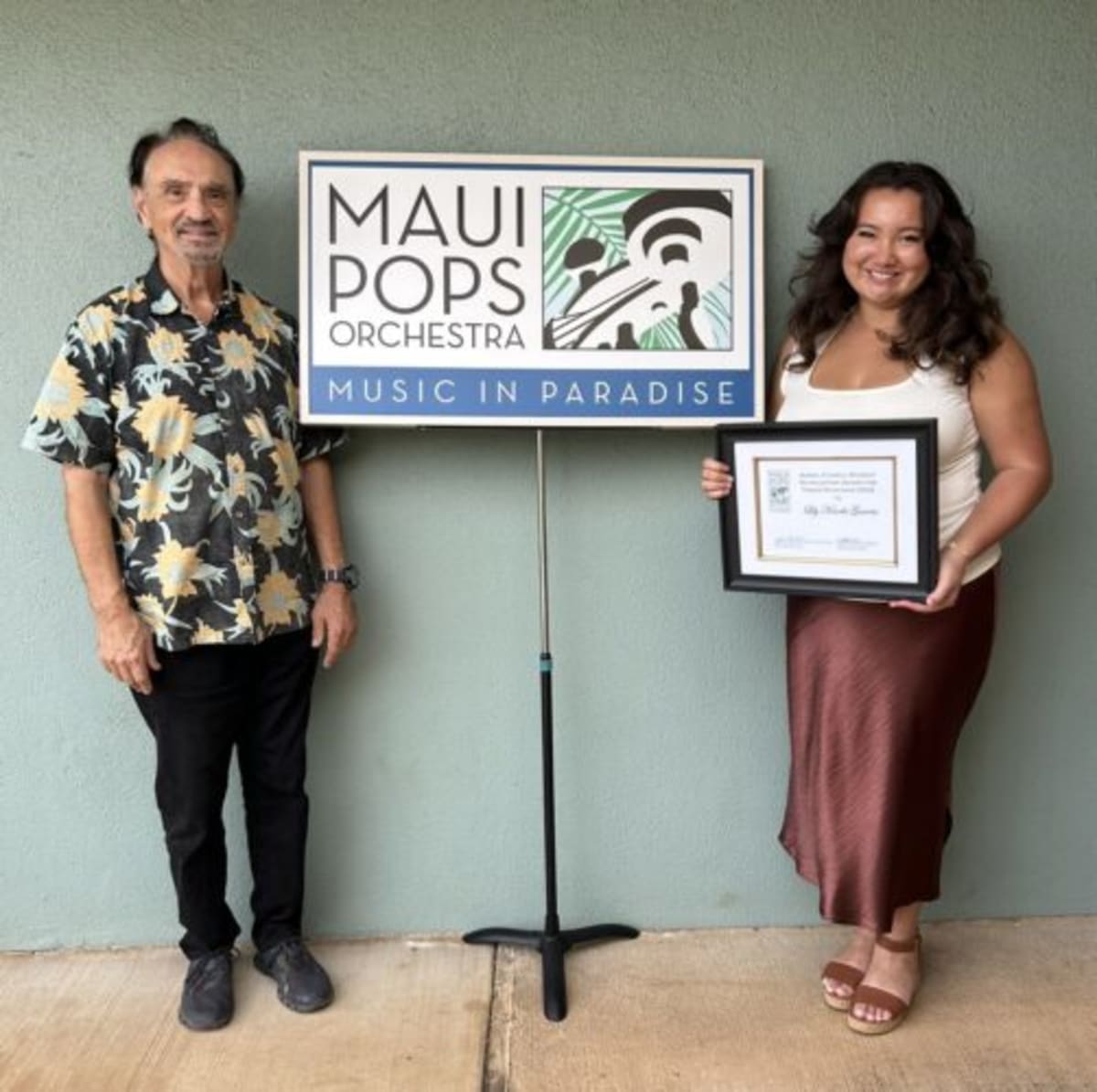 Young cellist Avery Pendergraft stands beside Maui Pops Orchestra conductor holding scholarship award certificate
