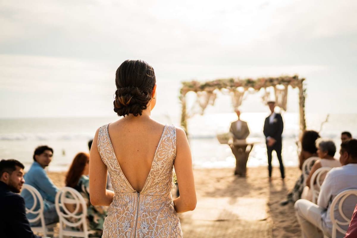 Bride in white dress walks on sandy beach toward ocean for wedding ceremony
