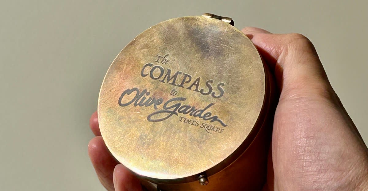 Hand holding circular compass device with arrow pointing toward Times Square Olive Garden