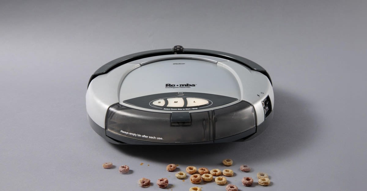 Early model Roomba robot vacuum cleaning a hardwood floor in a family home
