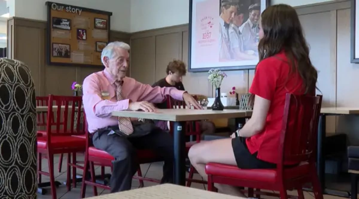 Smiling 92-year-old Gilbert Martin in Chick-fil-A uniform welcoming restaurant customers