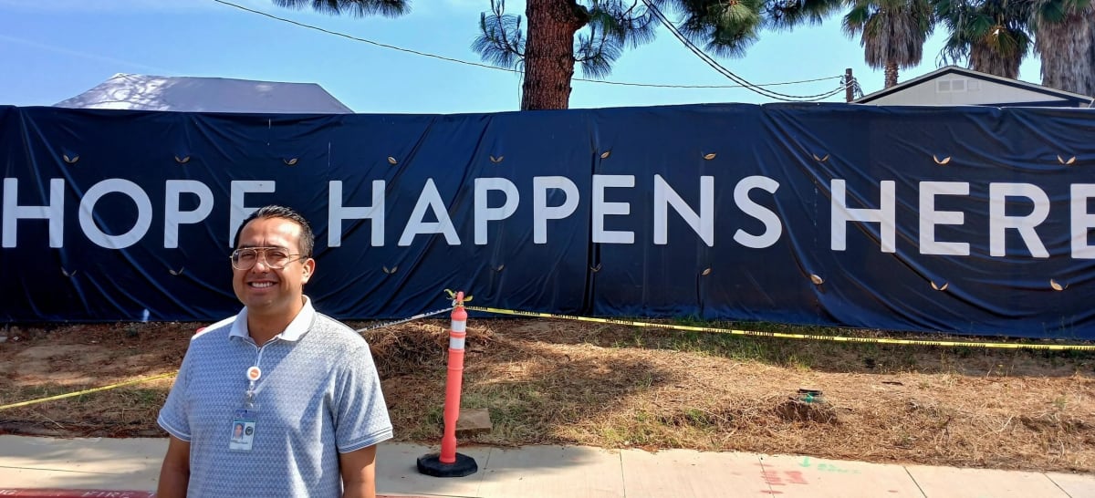 Man stands smiling in front of large banner reading Hope Happens Here at outdoor community event