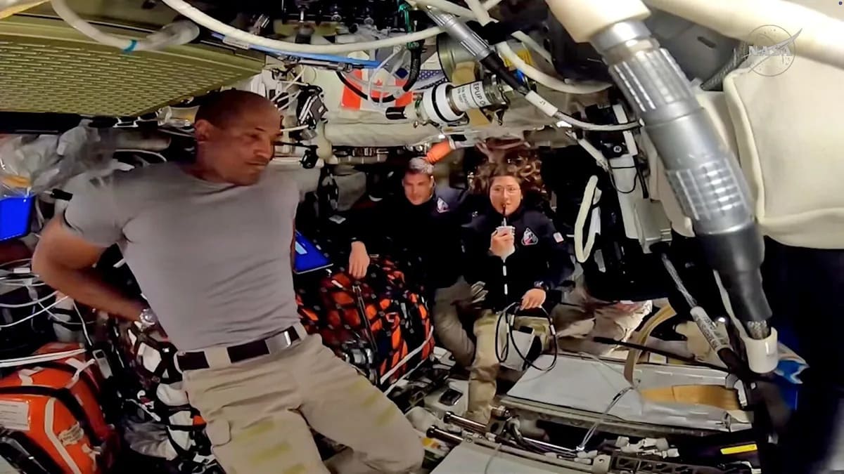 Three Artemis II astronauts floating inside Orion spacecraft waving at camera with Earth visible through window