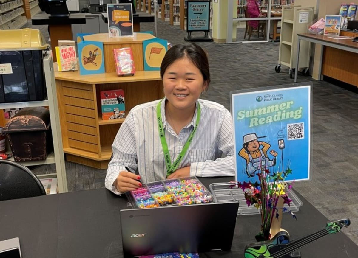** Teenage volunteer helping young child select books at colorful library summer reading program display