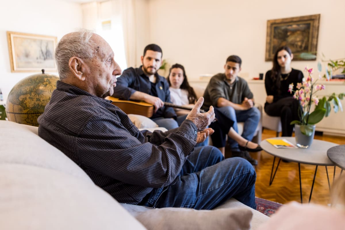 ** Holocaust survivor Tommy Shaham sharing testimony during intimate Zikaron BaSalon living room gathering