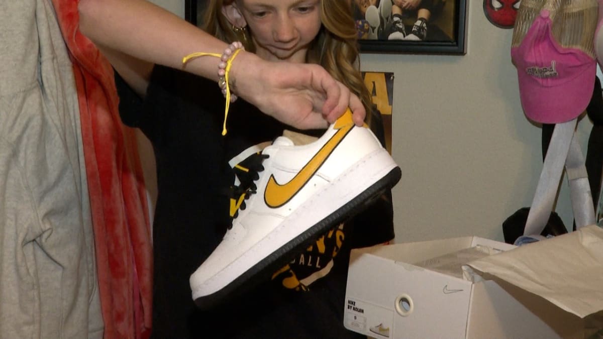 Fourteen-year-old Harper Stribe smiling while holding black and yellow Nike sneakers she designed for Iowa basketball