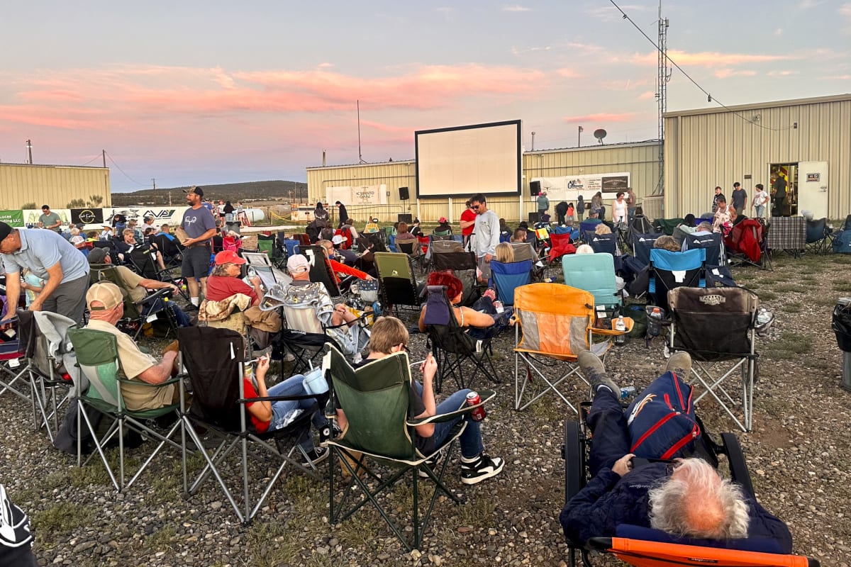 Families sitting on blankets in dirt lot watching outdoor movie screen at dusk in Colorado