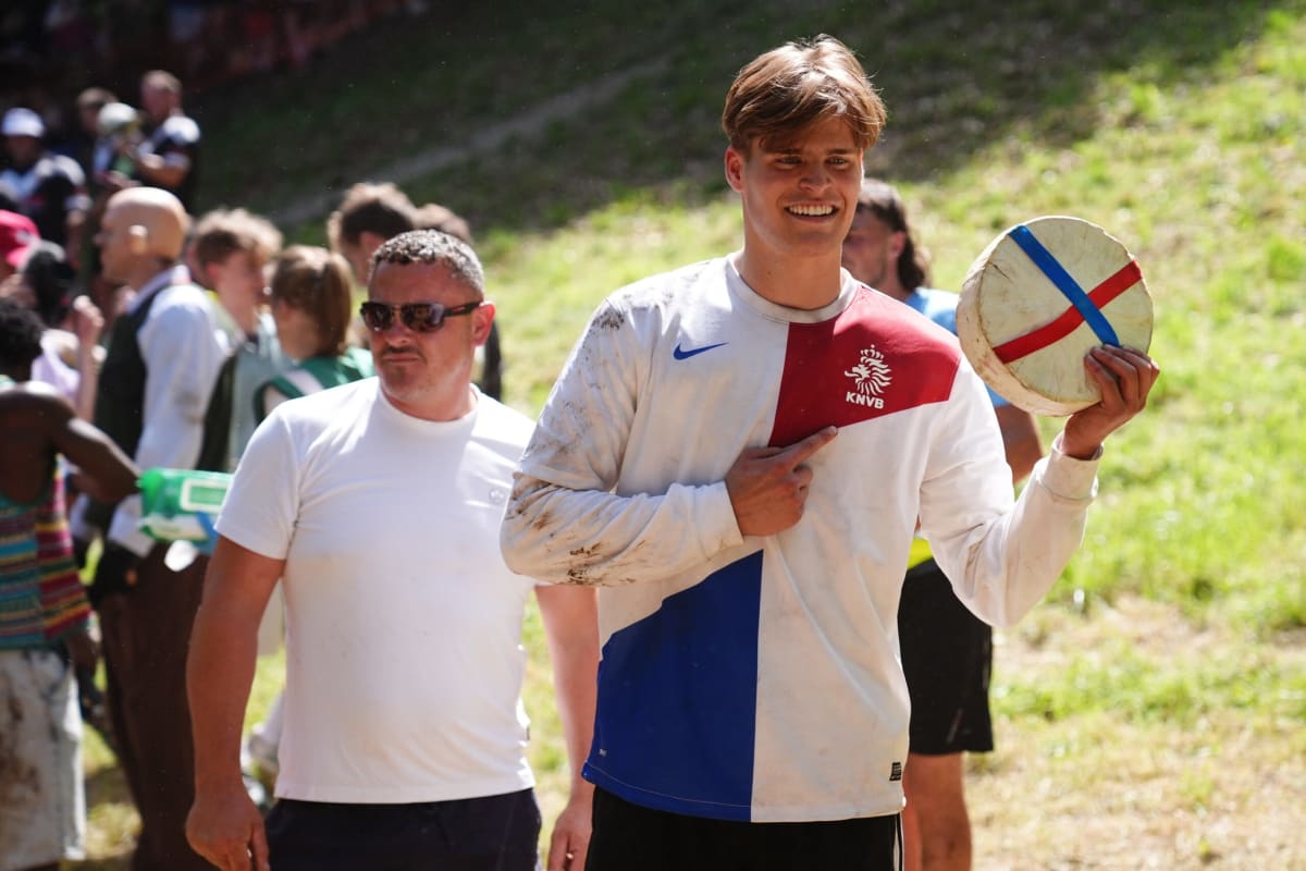 Young man in orange Dutch football shirt holds large round cheese wheel triumphantly above crowd
