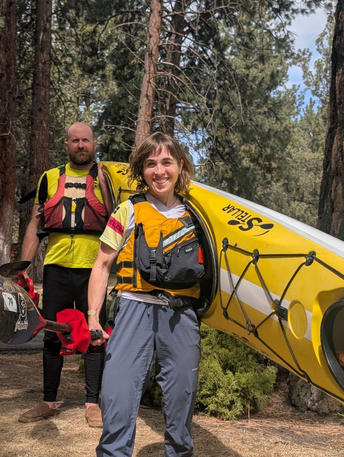 Two kayakers paddling down a scenic river with forested banks under clear skies