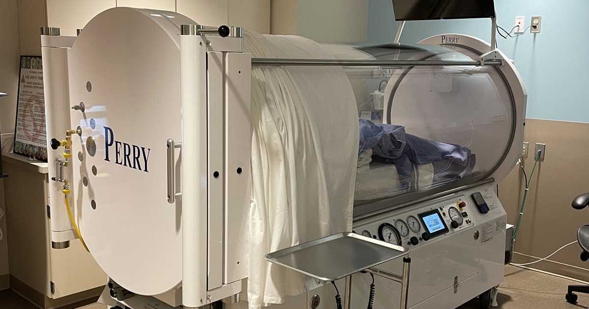 Smiling cancer survivor standing beside a clear hyperbaric oxygen chamber in hospital treatment room