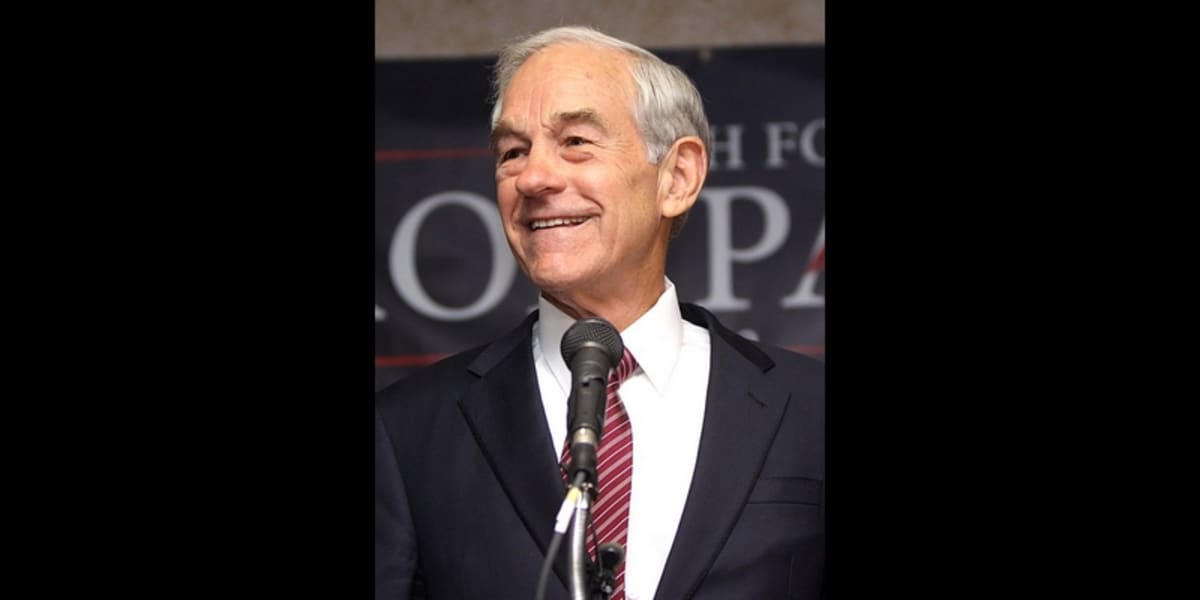 ** Former Congressman Ron Paul speaking at an event, smiling in a dark suit