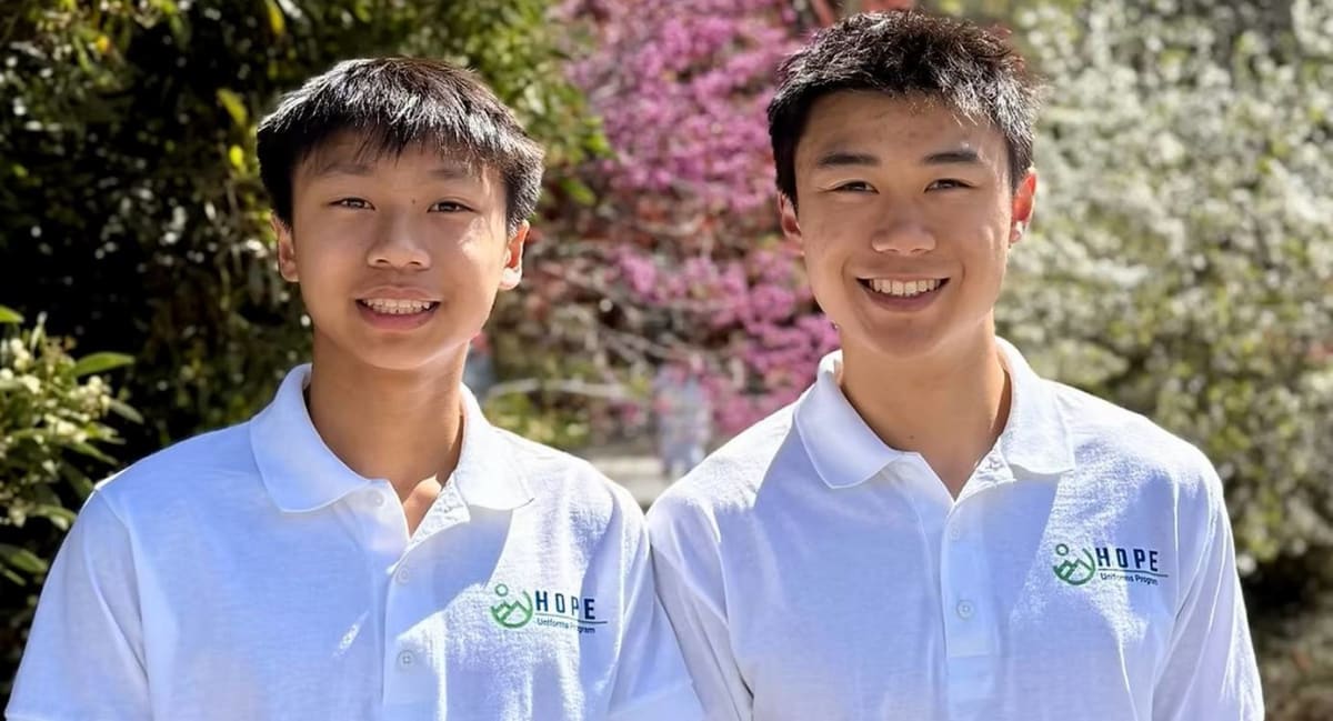 Teen brothers Desmond and Ethan Hua stand beside organized bins of donated school uniforms in garage