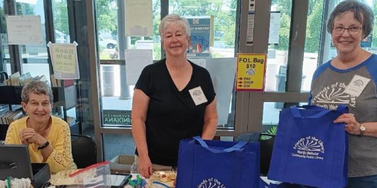 Three smiling volunteers stand behind tables of books at library book sale event
