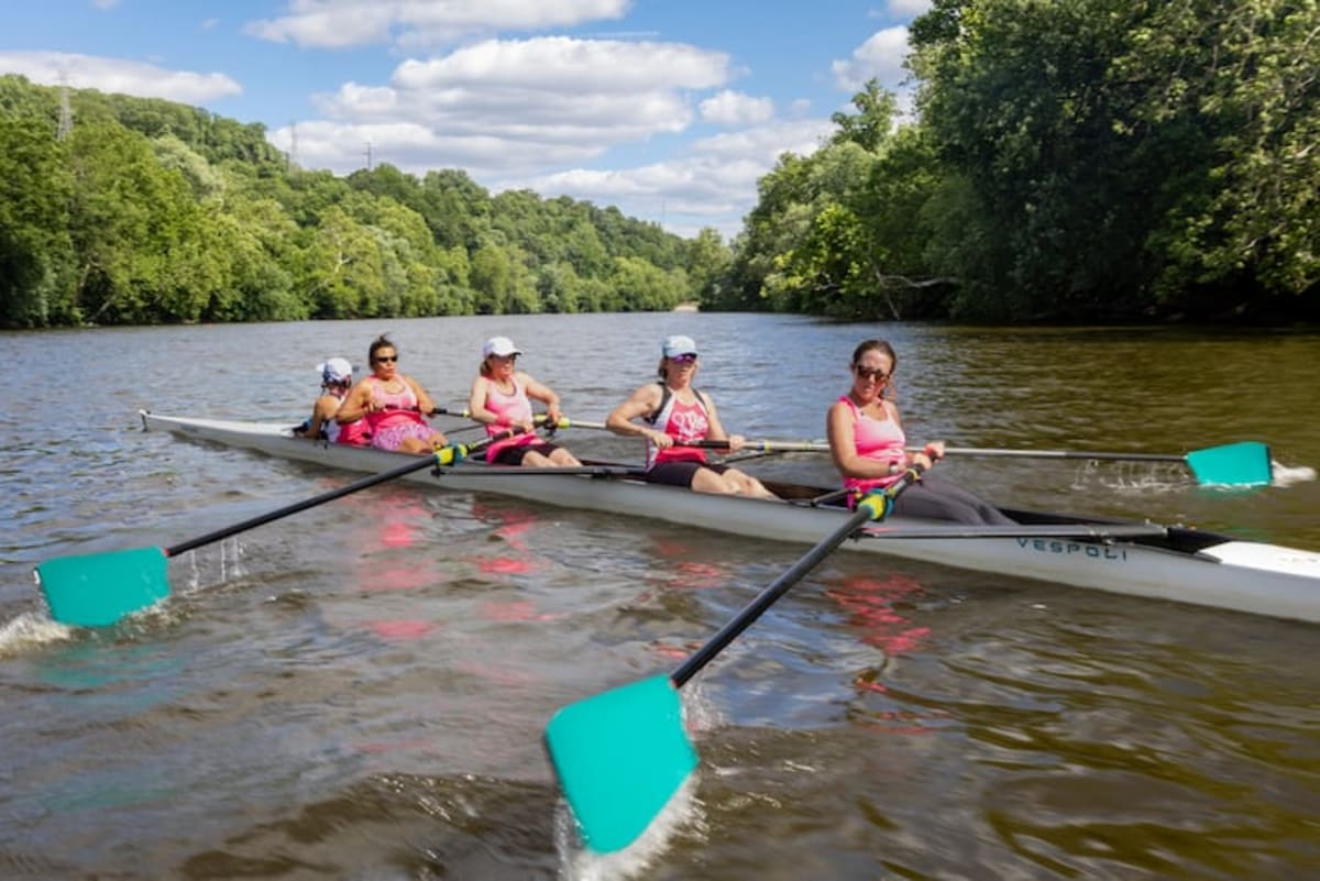 Five women breast cancer survivors in rowing gear smile together holding oars at boat dock