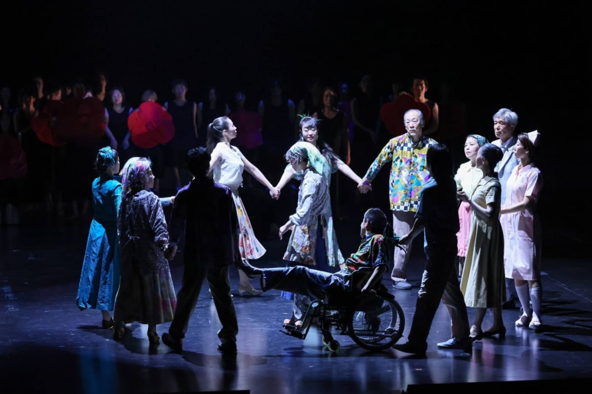Large diverse theater troupe performing on stage with colorful hanging garments overhead