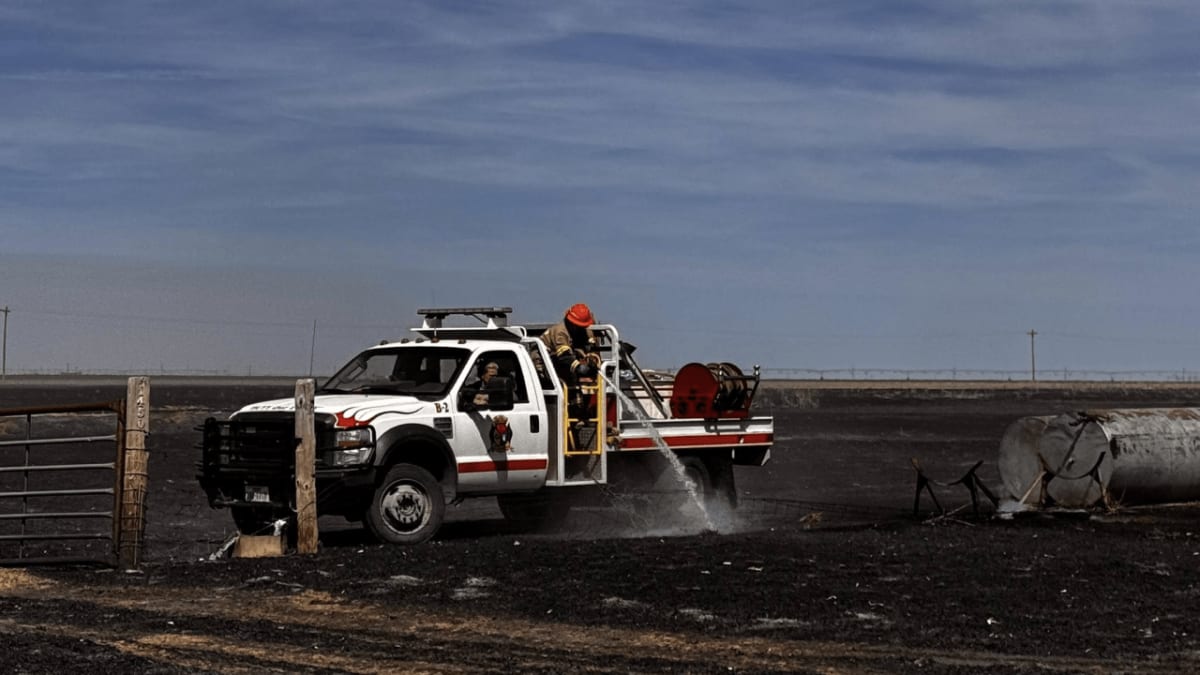 Firefighters battling brush fire with equipment near rural Texas structures under smoky conditions