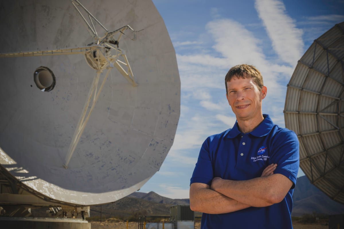 Erik Richards stands in front of large white satellite antennas at NASA's White Sands facility