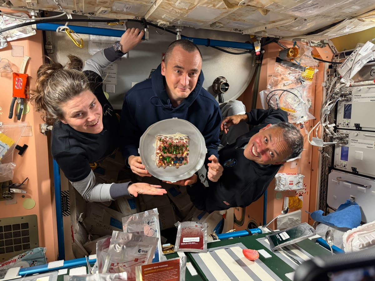 Three smiling astronauts in blue flight suits holding cake aboard International Space Station
