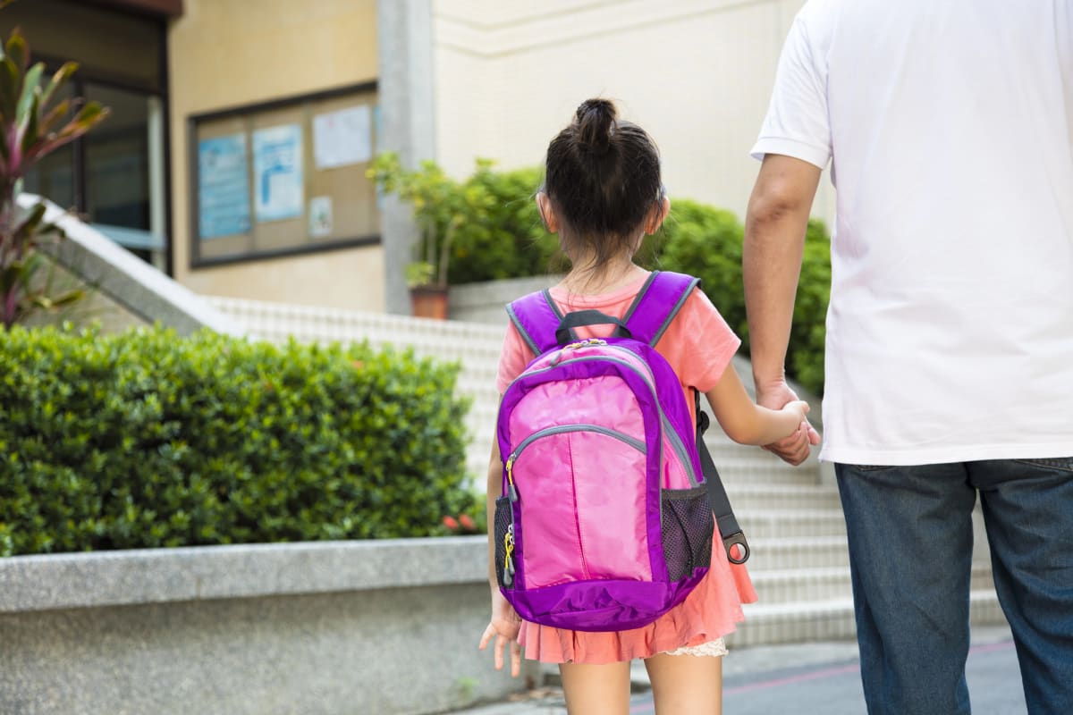 Parent walking young child with backpack toward school building on sunny morning