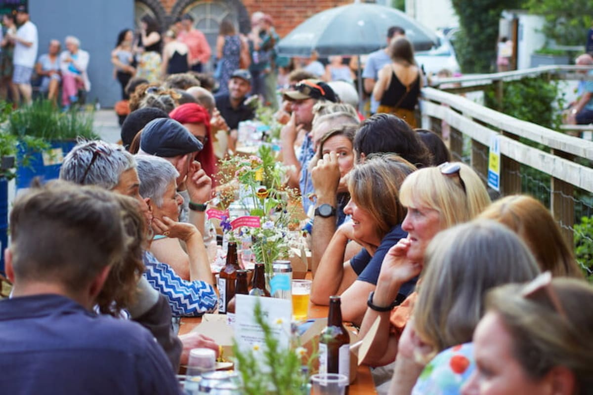 Diners of all ages sitting together at long wooden tables sharing a meal in rustic industrial space