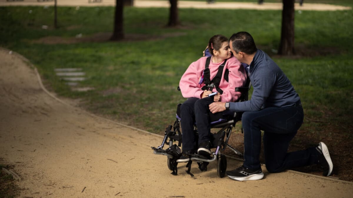 Father sitting beside teenage daughter with long braided hair in medical setting, showing devoted care