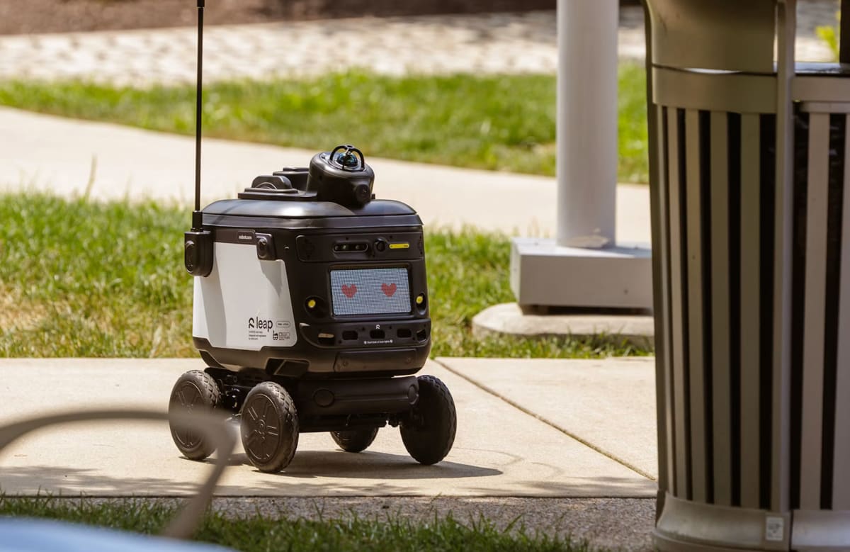 Small white delivery robot with Robot.com branding rolling on college campus sidewalk