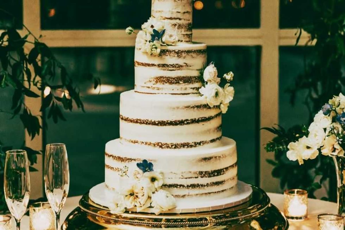Groom calmly gesturing toward fallen wedding cake on floor while bride watches with loving expression