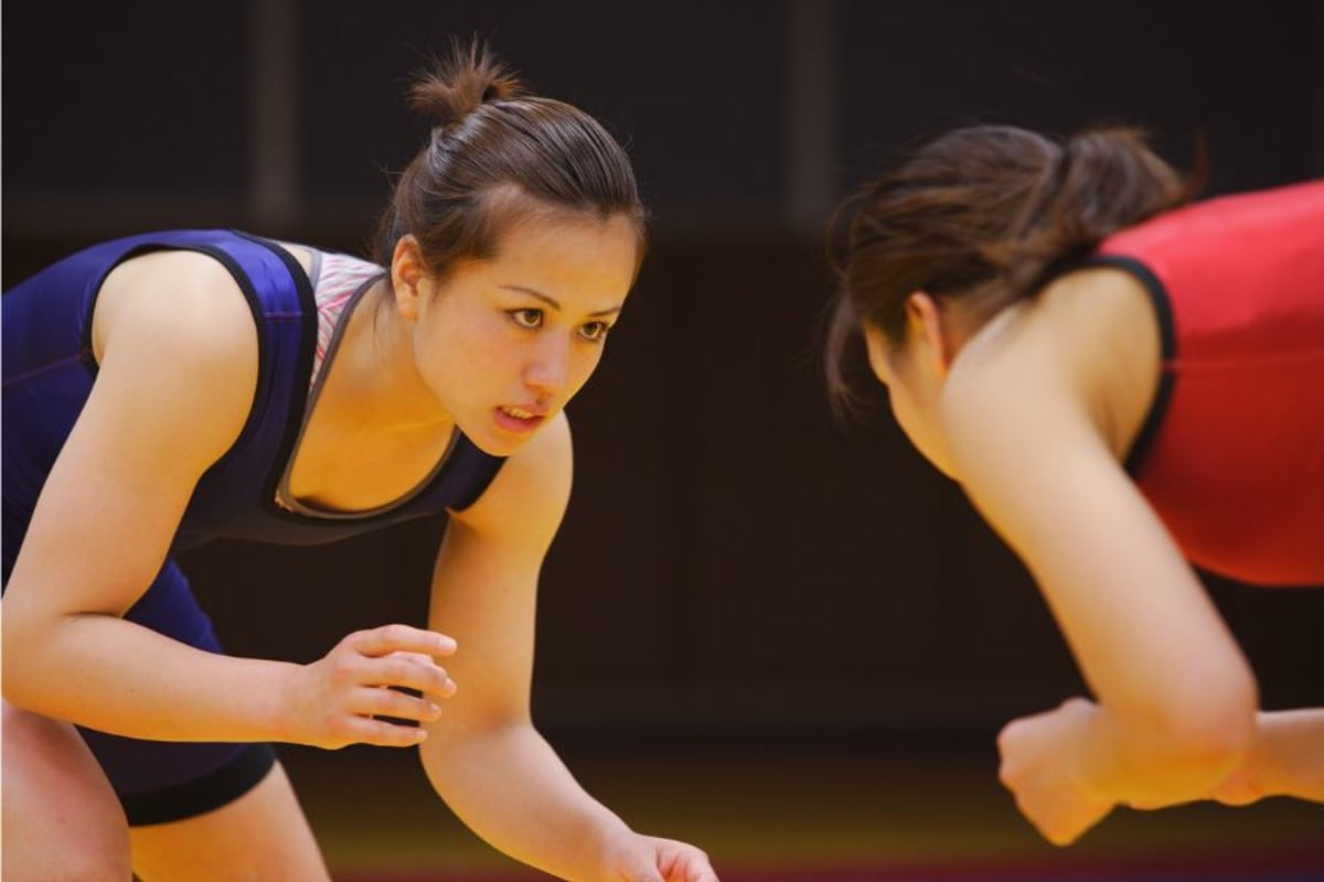 Female wrestler in blue singlet helping guide nervous opponent into proper wrestling stance
