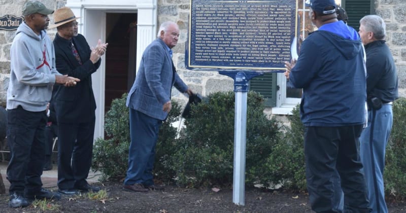 Community leaders unveiling historical memorial marker at ceremony, showing people gathered for reconciliation event in Maryland