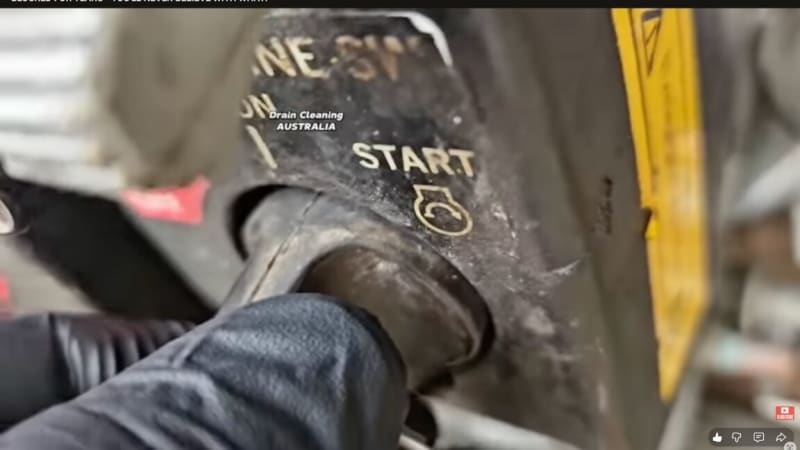 Plumber's gloved hands operating high-pressure water jet equipment over drainage system opening