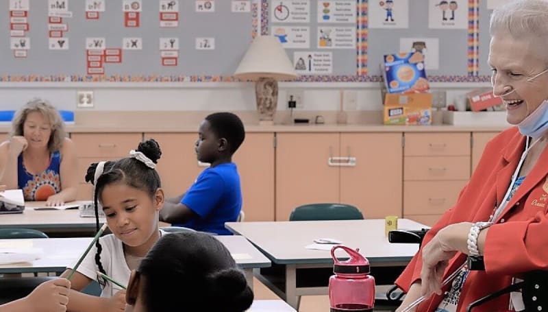 Senior volunteer sitting with elementary student reading book together in bright classroom