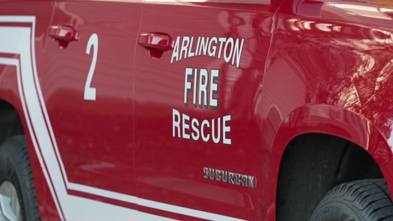 Arlington firefighter holding rescued dog outside smoke-damaged two-story home on sunny morning