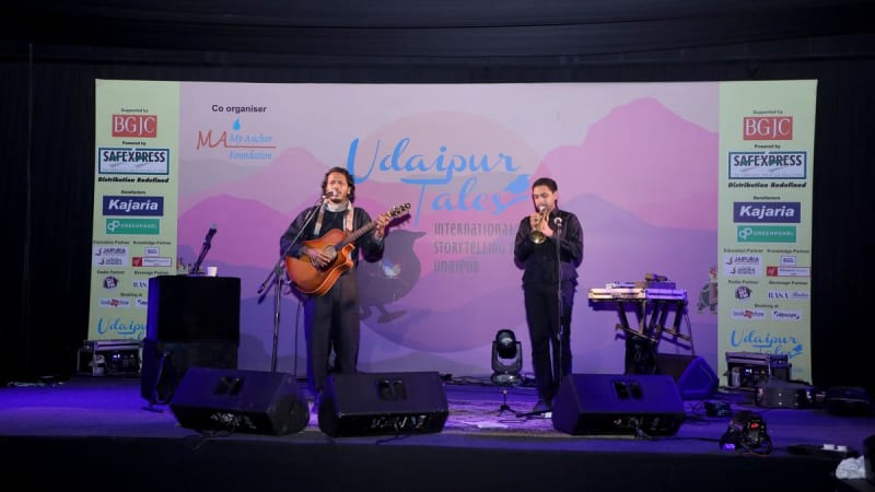 Diverse storytellers performing on stage at Udaipur Tales Festival with engaged audience listening attentively in warm lighting